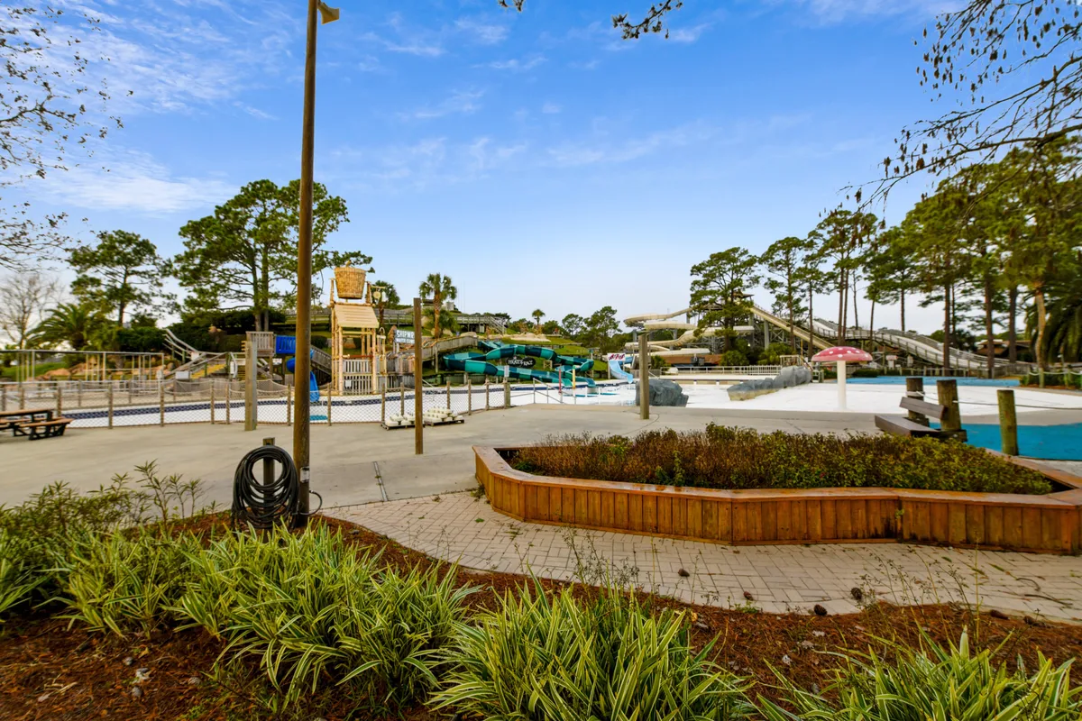 A lively resort pool area featuring water slides, a swimming pool, landscaped gardens, and families enjoying the amenities. The scene is bright and joyful, perfect for summer and spring activities.