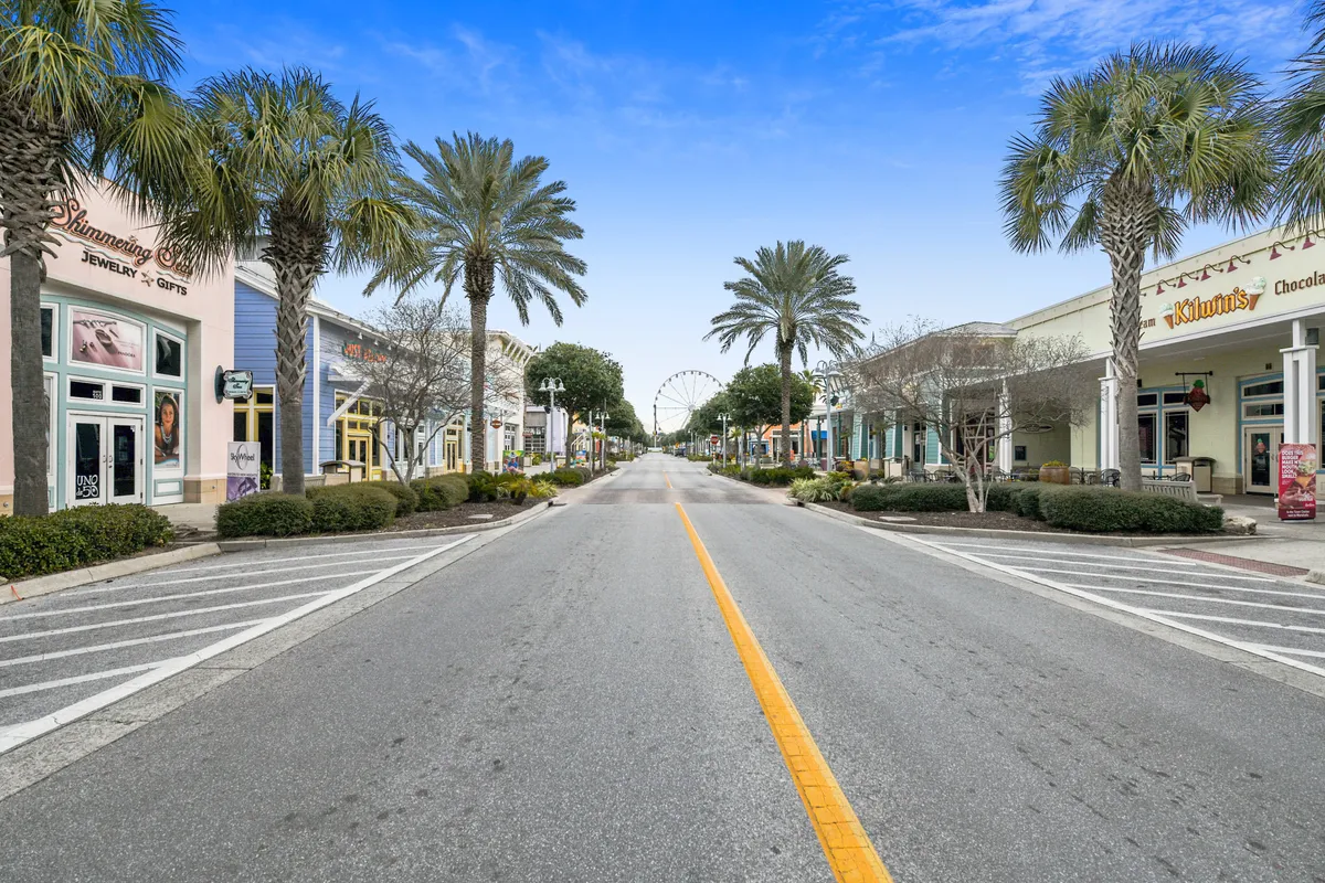 This vibrant exterior image shows palm trees framing a lively pedestrian promenade at Pier Park, with a Ferris wheel in the background and people enjoying shopping, dining, and strolling. The scene captures the essence of Pier Park’s bustling atmosphere and its role as a central hub for entertainment and leisure.