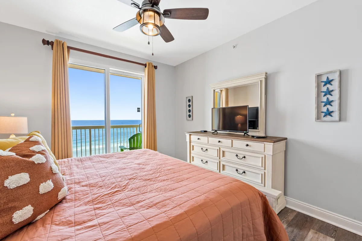 A serene bedroom featuring a bright orange quilt on a king-size bed, a large window framing expansive ocean views, and a wooden dresser topped with a TV. Sunlight pours into the room, creating a tranquil, restful vibe ideal for winding down after a sun-filled day at the beach.