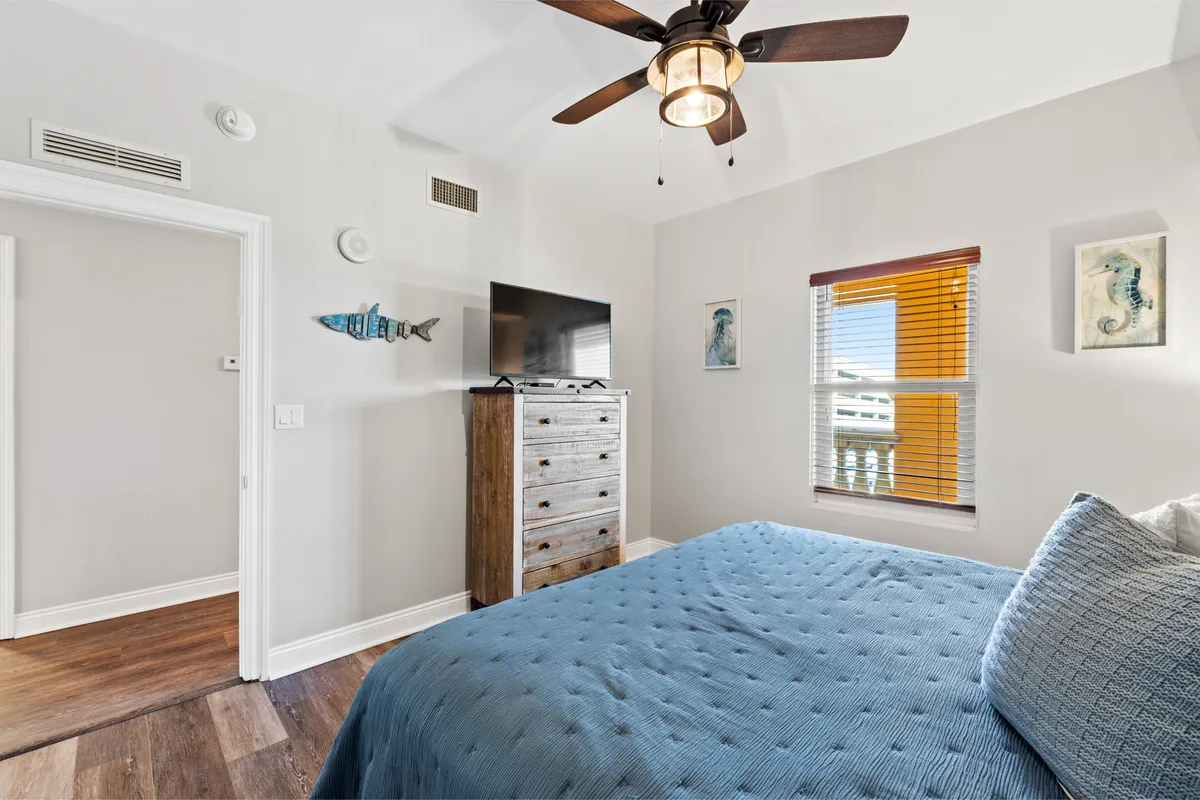 A cozy bedroom with a king bed adorned in textured bedding, a wooden dresser, television, window with blinds, and tasteful wall art. The room is filled with soft, natural light, inviting guests to unwind and relax in a comfortable, private space.