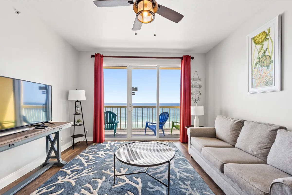 A bright, open living room featuring a plush sofa and coffee table, flat screen television, and large sliding doors leading out to a private balcony. The space is oriented toward expansive ocean views and balcony seating, inviting natural light and a sense of openness.