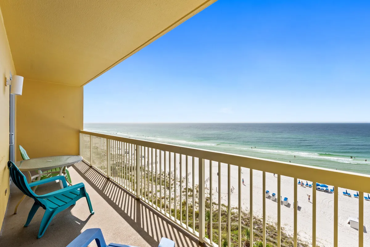 A private balcony with comfortable seating and a small table, offering a panoramic view of the white sand beach and turquoise Gulf waters beneath a clear blue sky. Shadows from the balcony railing create geometric patterns on the floor, enhancing the sense of relaxation and tranquility.