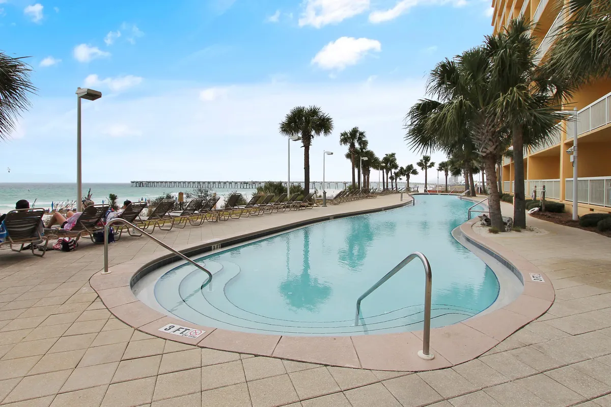 A curved swimming pool surrounded by rows of lounge chairs and lined with palm trees, with a view of the pier and Gulf waters in the distance. The pool area appears inviting, sunlit, and part of a well-maintained resort setting.