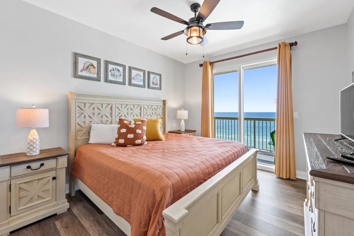 A spacious primary bedroom featuring a king size bed with a coral-colored quilt, a vintage-style dresser with a flat-screen TV, a ceiling fan, and a balcony door offering a stunning gulf view. Natural light floods the room, creating a serene and inviting atmosphere.