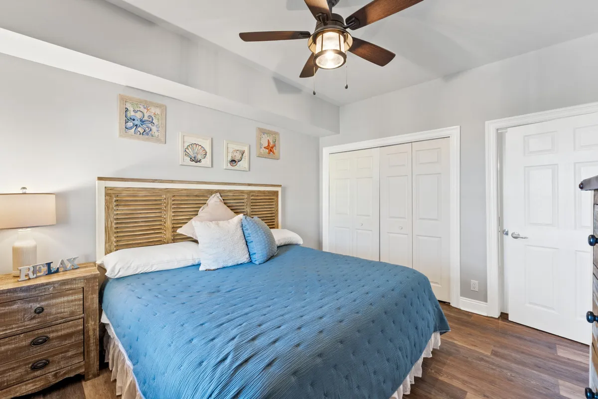 A peaceful second bedroom with a king size bed dressed in soft blue bedding, a wooden headboard, matching nightstand, ceiling fan, and wall art depicting seashells and starfish. The space exudes a calm, relaxing vibe, perfect for restful nights.