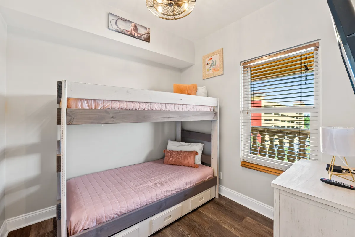 A cheerful bunk bed alcove with pastel pink bedding, a window with wooden blinds offering a glimpse of the balcony and resort exterior, a white dresser with a lamp, and playful beach-themed wall art. The space is bright and ideal for children or teens.