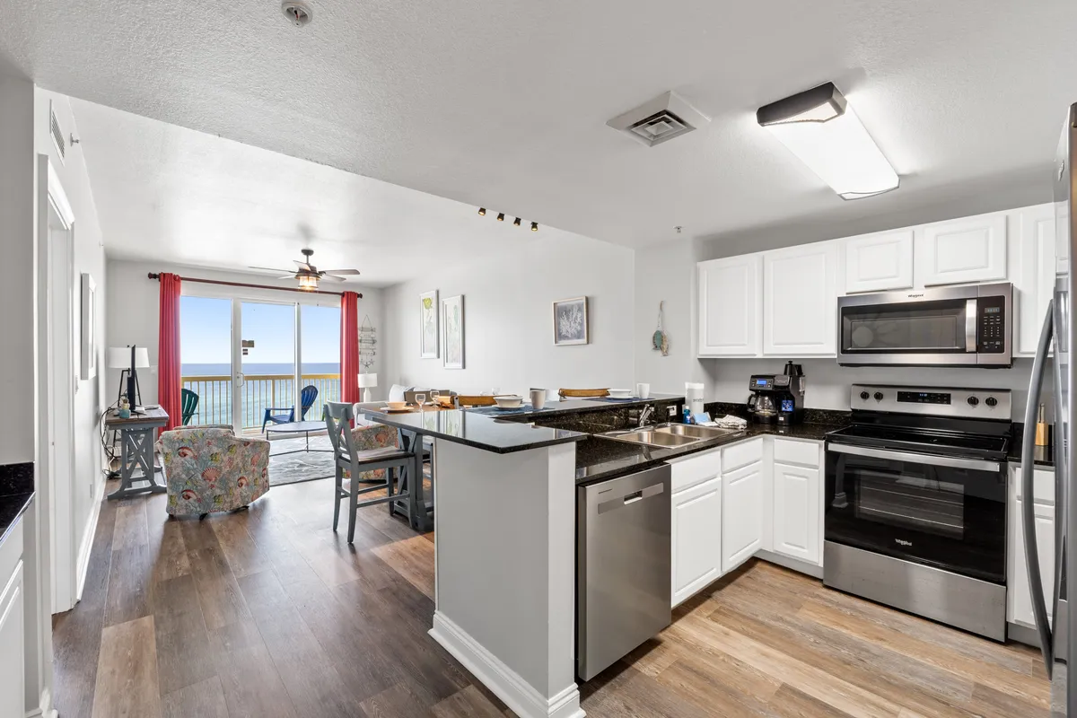 A bright, open space combining a kitchen with a granite-topped island, a dining table elegantly set for six, and a cozy living area with a colorful armchair. The balcony, visible through the glass doors, offers panoramic ocean views and is furnished for outdoor enjoyment.
