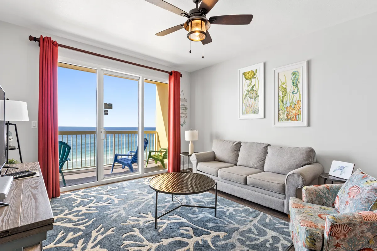 A spacious living area featuring a gray sleeper sofa, a colorful floral armchair, and a round coffee table. Large sliding glass doors open directly onto a private balcony with sweeping Gulf ocean views, inviting natural light and the scenery inside.