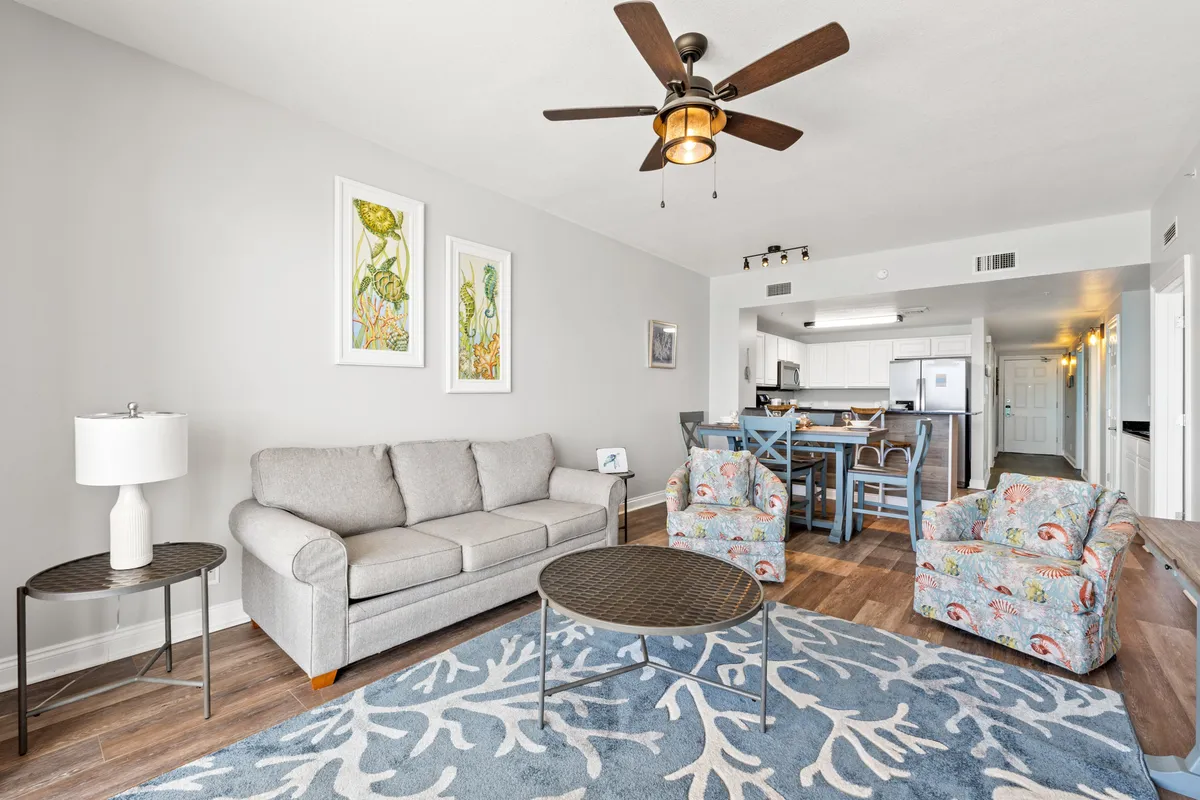 This image presents a bright, inviting living area with a light gray sofa, floral patterned armchairs, a round metal coffee table, and a blue coral-patterned rug. The space is open and airy, hinting at the comfort and relaxation families can expect indoors, with easy access to amenities and plenty of room for gathering after a day of adventure.