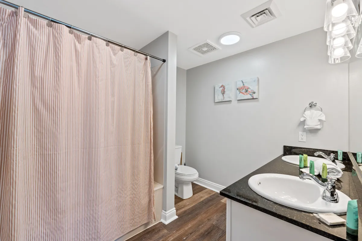 A bathroom scene featuring a double sink vanity with a black countertop, a partially visible toilet area, a striped shower curtain, and decorative flamingo artwork on the wall. The space is fresh and functional, perfect for families getting ready for local adventures or winding down after a busy day.
