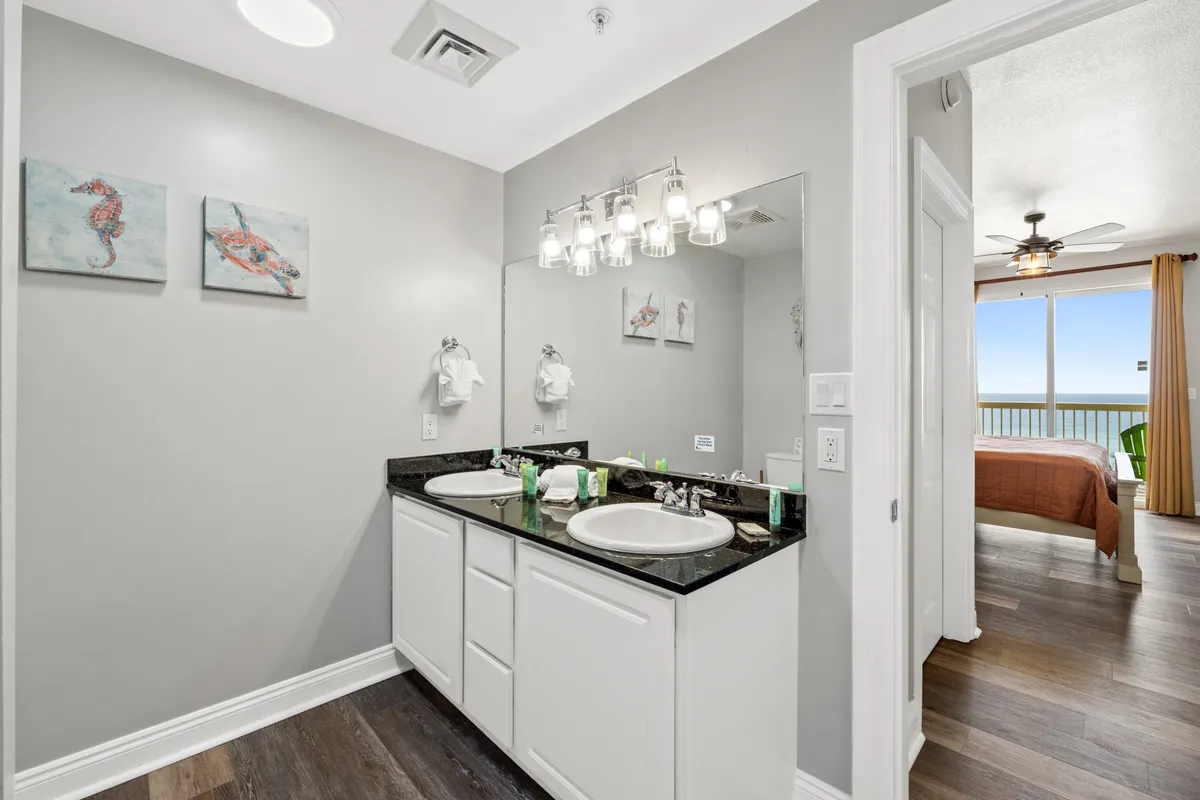 A bright bathroom with a dual sink vanity and large mirror, decorated with coastal artwork. There's a view into the primary bedroom with a king size bed and balcony beyond, illuminated by natural light. The bathroom is clean, modern, and connected to the sleeping areas.
