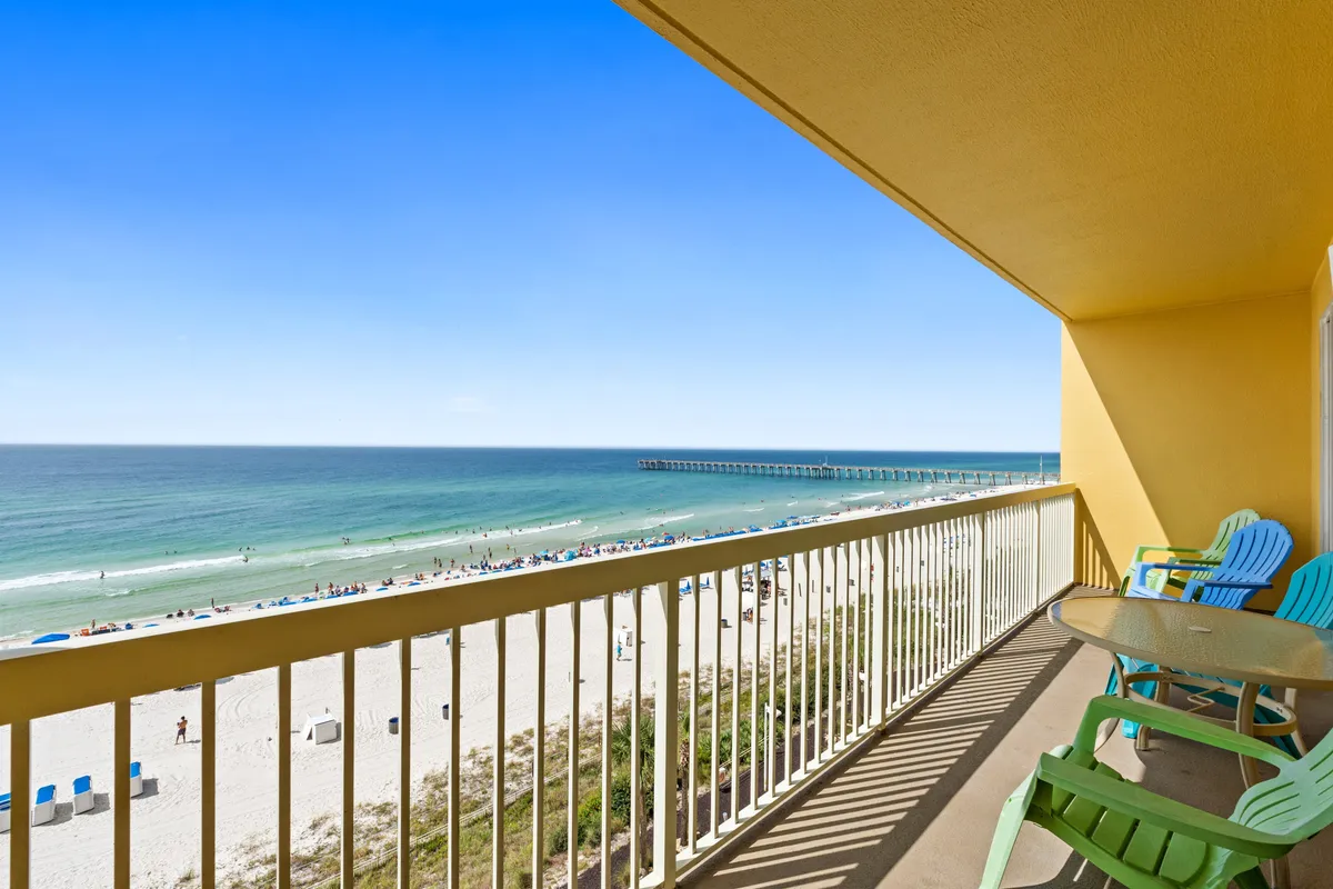 A furnished balcony with colorful chairs and a table overlooks an expansive white sandy beach, blue ocean waves, and a long pier stretching into the Gulf. The scene is peaceful yet vibrant, inviting moments of relaxation, sunset watching, and connection to the beachfront environment.