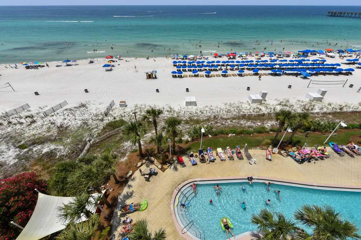 A vibrant exterior scene featuring a large swimming pool surrounded by palm trees, with people enjoying the water. In the background, there is a white sandy beach lined with umbrellas and loungers, and the turquoise Gulf waters where swimmers and boats are visible. The overall atmosphere is lively and inviting, capturing both poolside and beachfront resort experiences.