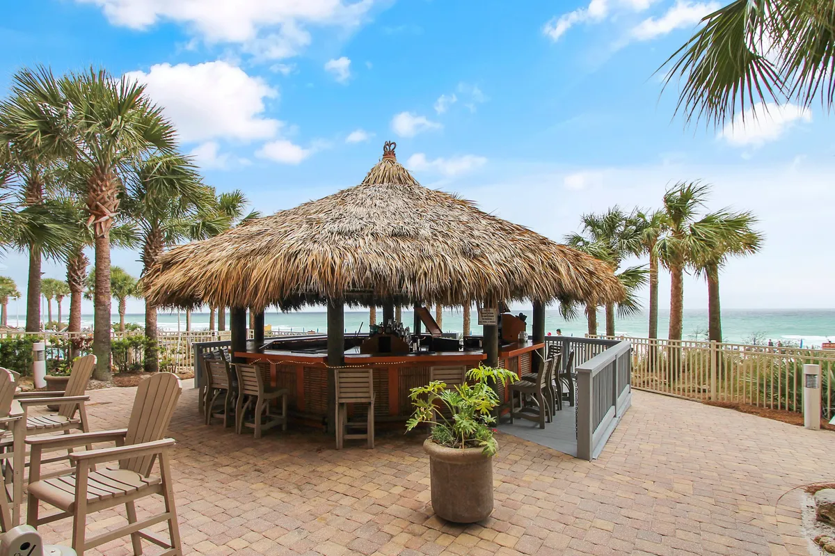 A thatched-roof tiki bar with seating, surrounded by palm trees and with an ocean view beyond a fence. Lounge chairs are set out on the paved patio, creating a festive and tropical atmosphere.