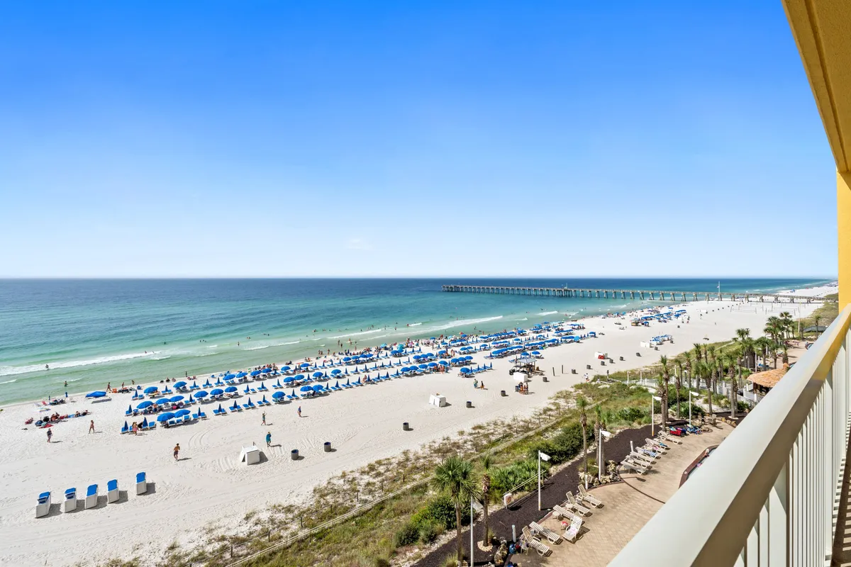 A stunning view from a condo balcony, overlooking a white sandy beach lined with blue umbrellas and lounge chairs. The calm turquoise ocean stretches out to the horizon, with a long pier extending into the Gulf and a hint of resort buildings in the distance. The scene is lively with families and groups enjoying the sun and surf.
