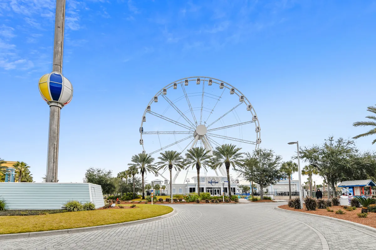 A cheerful scene featuring the iconic SkyWheel ferris wheel against a clear blue sky. The foreground includes palm trees, a paved walking path, and a whimsical balloon-shaped sculpture, with landscaped greenery inviting families to stroll, play, or relax.