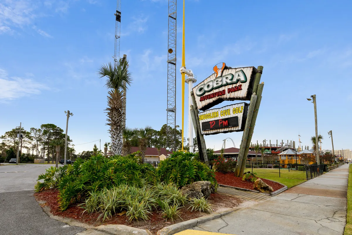 A vibrant exterior view of Cobra Adventure Park, with its colorful sign and towering amusement rides set among tropical landscaping. The park’s energetic atmosphere is evident, inviting families to explore thrilling rides and playful activities nearby.