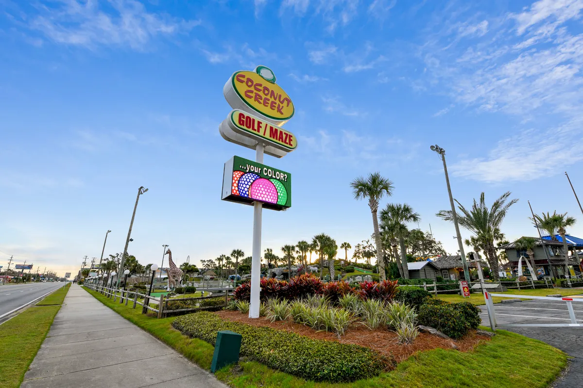 The Coconut Creek Golf / Maze sign stands out, surrounded by palm trees and whimsical tropical landscaping. Colorful digital displays and playful miniature golf structures are visible, drawing attention to fun group activities. The area is bustling with families and groups enjoying mini golf and navigating the maze, embodying lighthearted vacation fun.