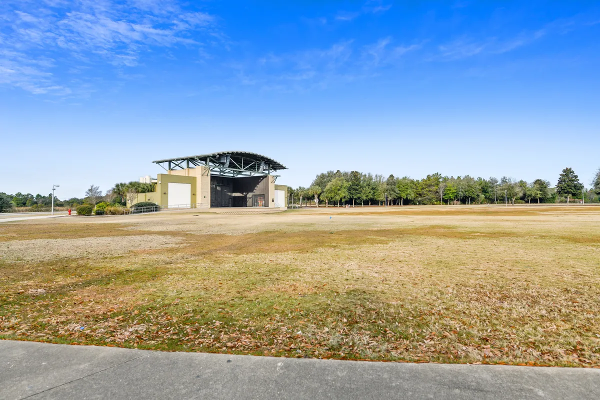 This image captures a wide open grassy field with a large covered pavilion or stage structure, set against a line of trees and a clear blue sky. The scene is peaceful but hints at the potential for outdoor festivals, concerts, and community events, with space for picnicking and gathering.