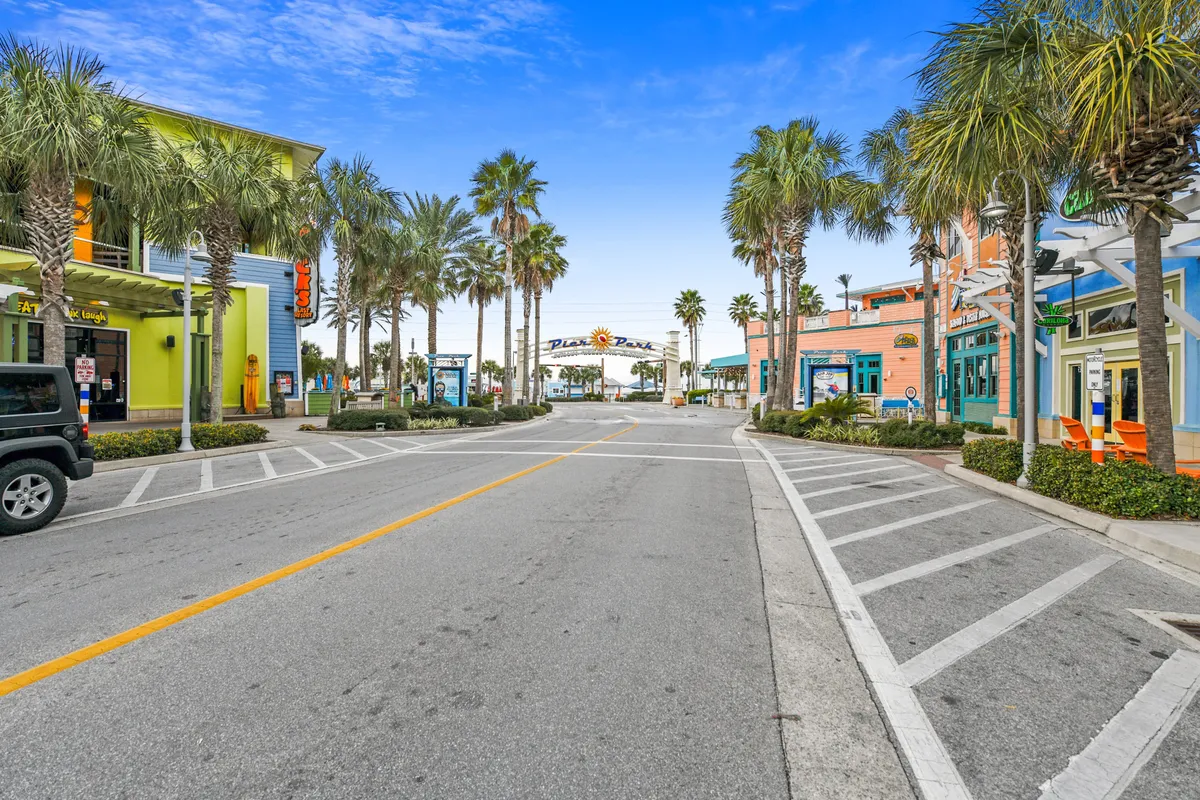 A lively street view of the Pier Park entrance sign, surrounded by towering palm trees and colorful building facades. Outdoor seating areas invite visitors to relax, while the street buzzes with the promise of diverse shopping, dining, and entertainment options.