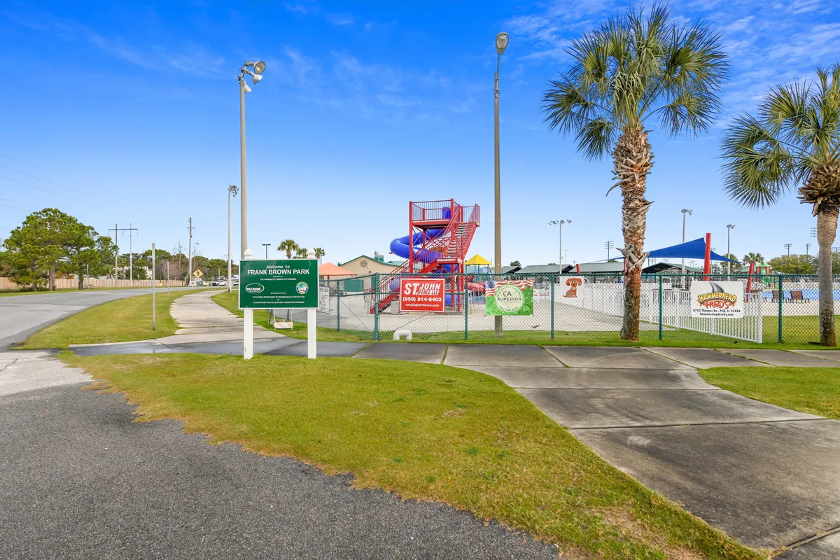 An energetic exterior shot of a waterpark area featuring colorful water slides, a fenced pool area with shaded seating, and palm trees nearby. The Frank Brown Park entrance sign is visible, suggesting proximity to family-friendly local activities and outdoor fun.