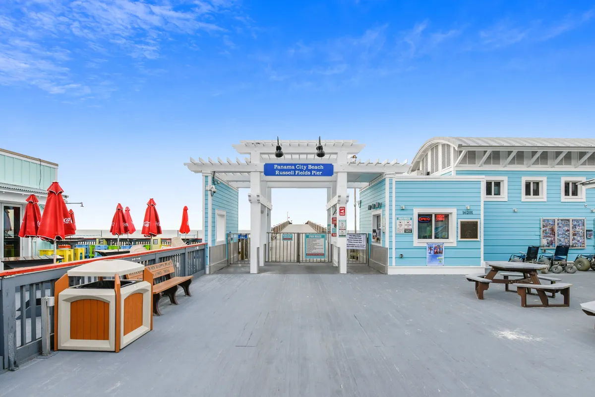 The entrance archway to Panama City Beach Russell Fields Pier is front and center, with clear signage welcoming guests. To the right, a light blue building with white trim adds a lively coastal touch, while red umbrellas and an outdoor seating area to the left invite visitors to relax. Benches and picnic tables dot the pier deck, and people are seen walking, fishing, and socializing, all with the ocean as a scenic backdrop.