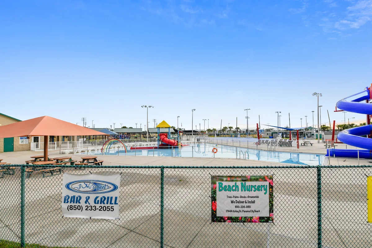 This lively exterior scene showcases a large swimming pool with water slides and splash features, shaded picnic tables under umbrellas, a fenced-in area for safety, and sports courts in the background. The environment is vibrant and inviting for families and groups, with a clear emphasis on fun, safety, and community.