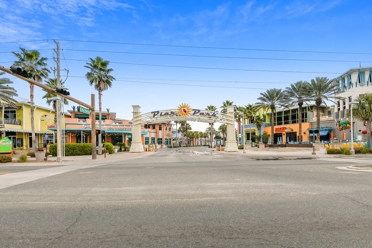 The Pier Park entrance arch welcomes visitors, with colorful storefronts lining both sides of a palm-tree-lined street under a clear blue sky. The area feels lively and festive, ideal for shopping, dining, and attending seasonal events.